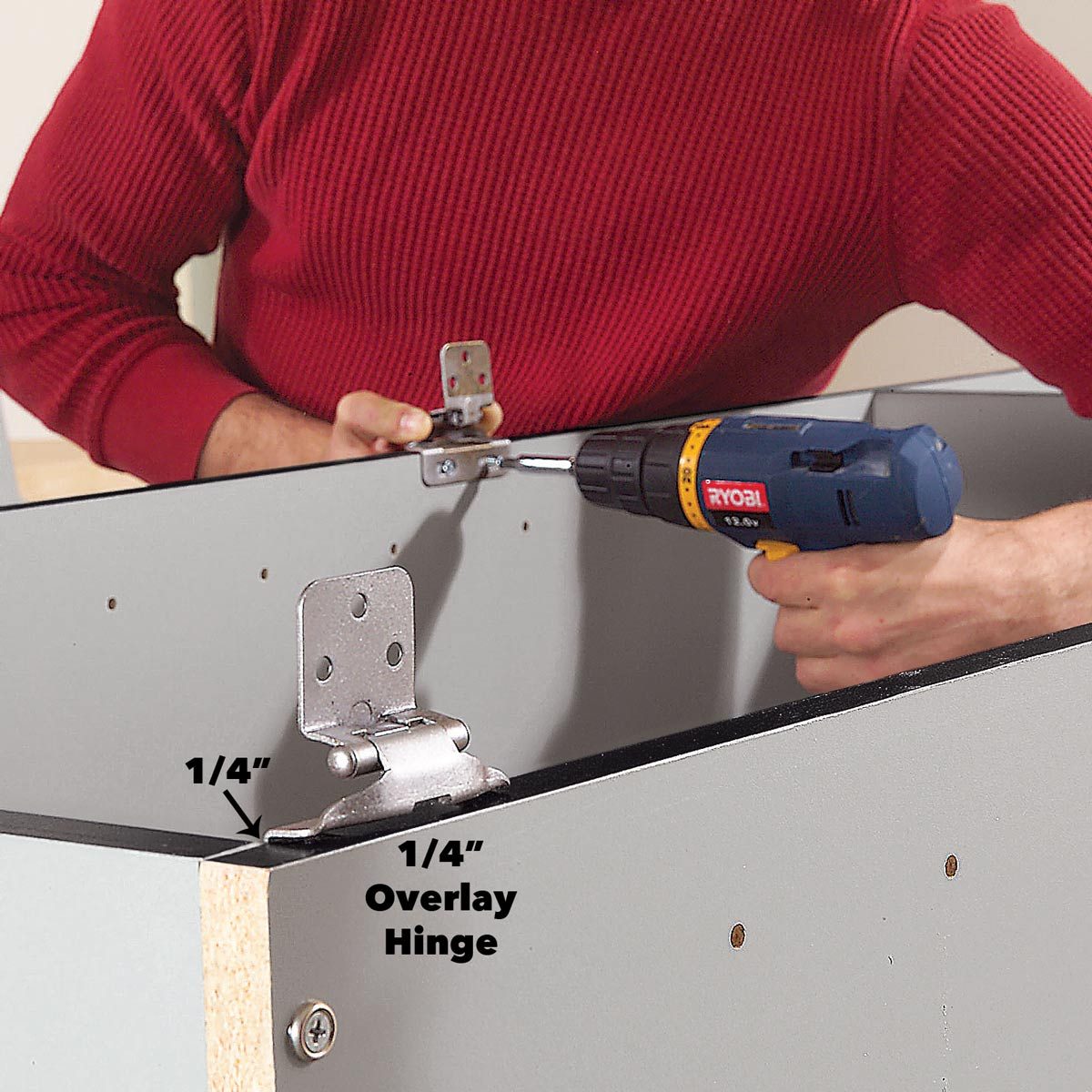A person is installing an overlay hinge onto a cabinet using a power drill, surrounded by a gray cabinet structure with visible pre-drilled holes.