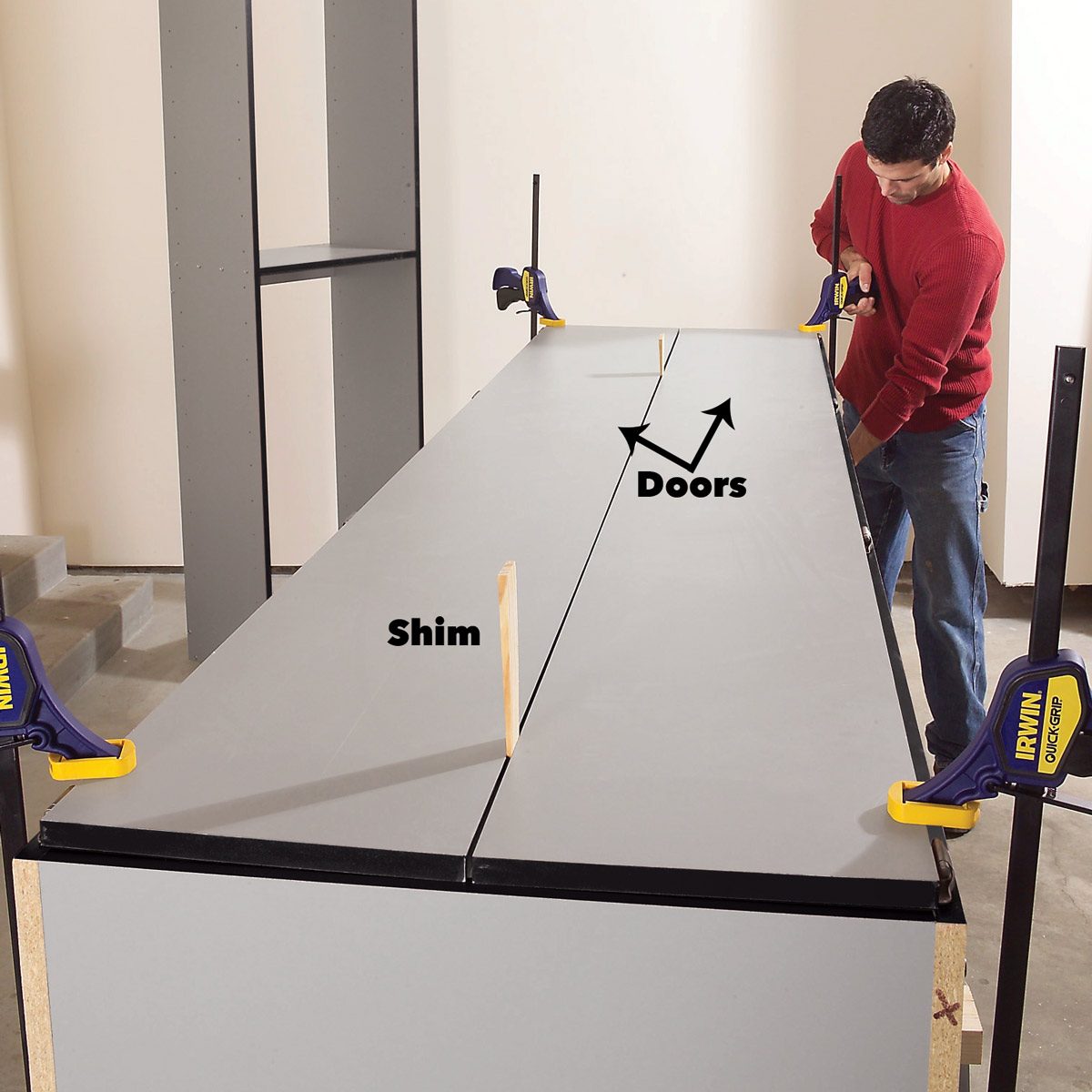 A man adjusts two gray boards while using clamps and a shim to align them, preparing for door installation in a neutral-colored room.