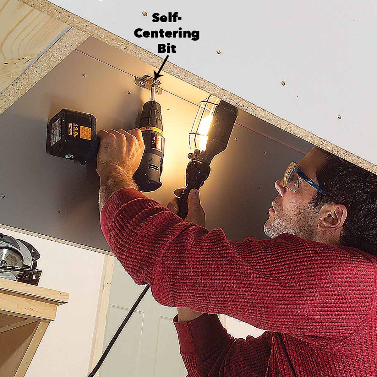 A person in glasses drills into a surface using a self-centering bit, while holding a light source, in a workshop setting.