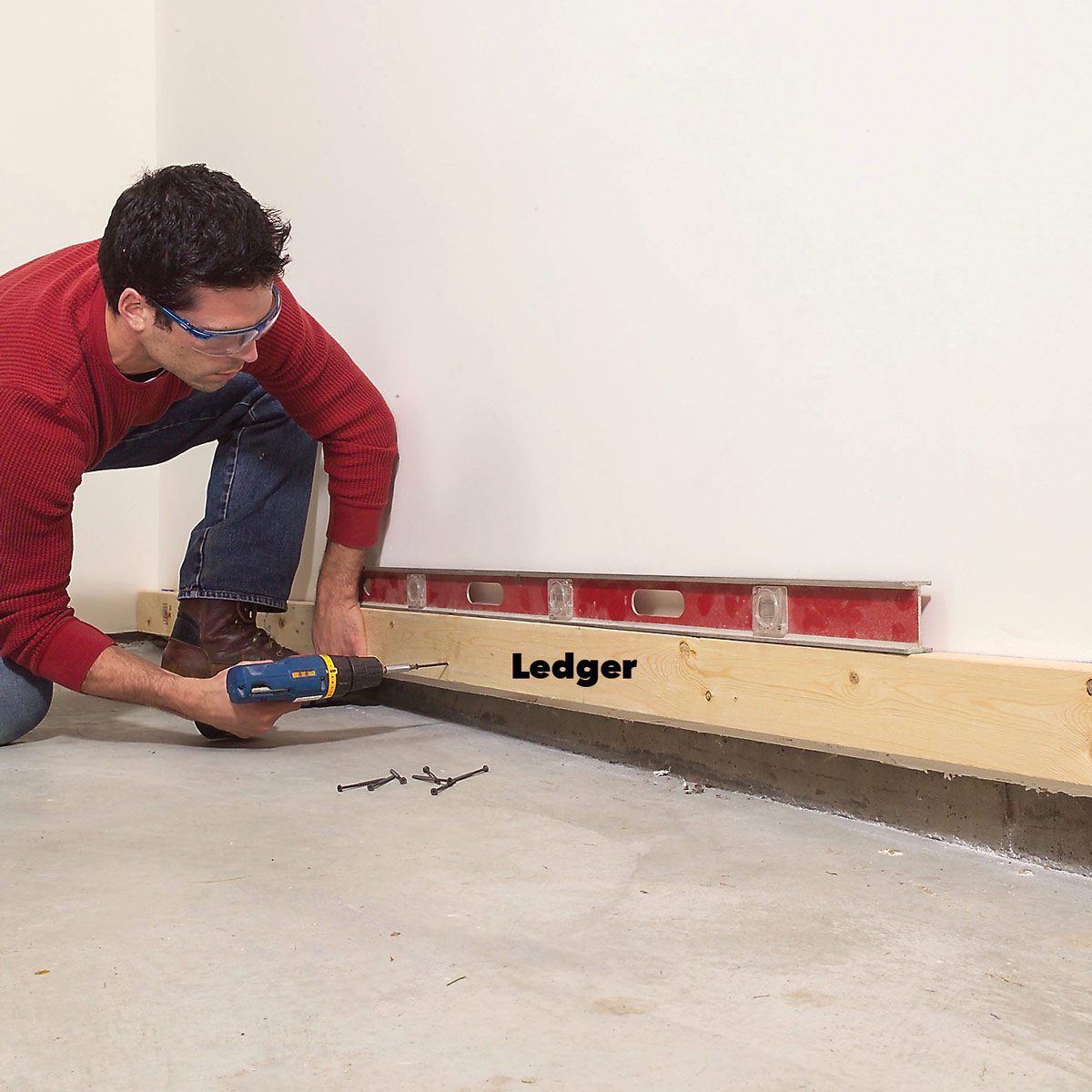 A man is drilling into a wooden ledger board attached to a wall on a concrete floor, using a level for accuracy.