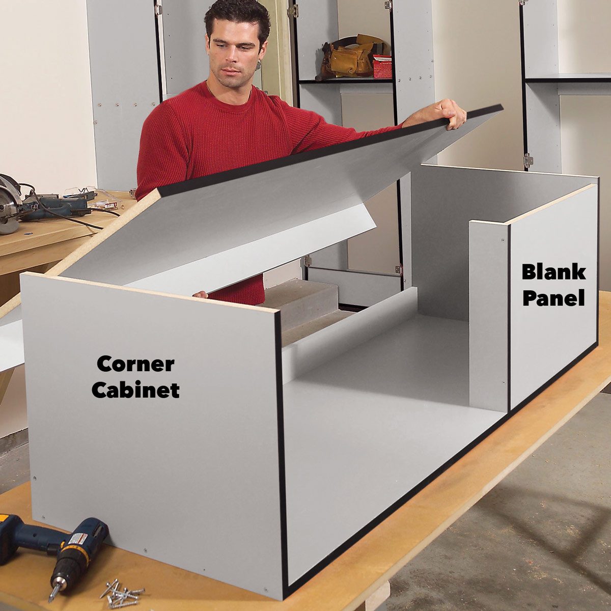 A man lifts the top of a gray corner cabinet, revealing its interior components and a blank panel, while surrounded by woodworking tools in a workshop setting.