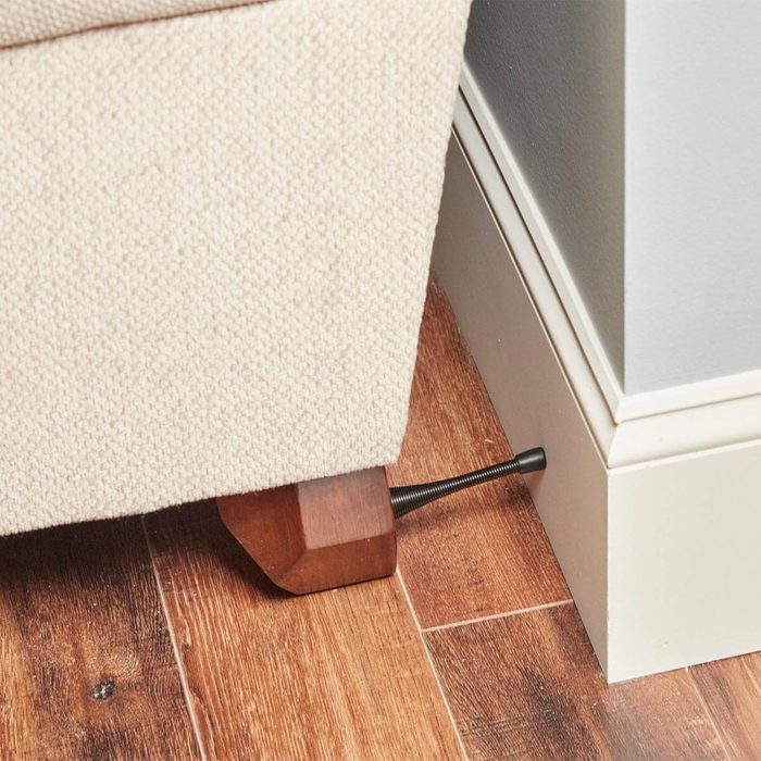 A couch leg is positioned on a wooden floor, with a metal screw adjusting its height against a wall's baseboard.