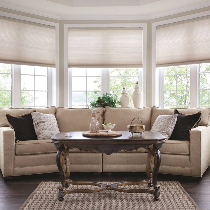 A cream-colored sectional sofa sits in a bright room, accompanied by a dark wooden table and decorative items, with large windows letting in natural light.