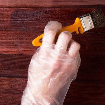 A gloved hand holds a yellow-handled paintbrush, applying finish to a wooden surface with rich brown tones.
