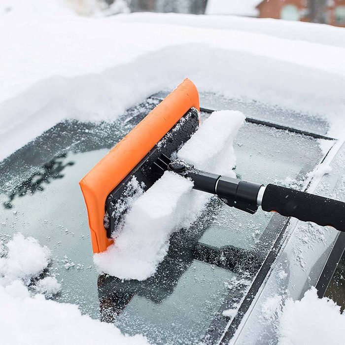 A snow brush clears a layer of snow from a car's roof. The surrounding area is covered in fresh, white snow, indicating winter conditions.