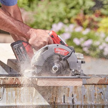 A circular saw cuts through a wooden plank, creating sawdust, while a person's hand grips the tool in a garden-like setting with blurred greenery and flowers.