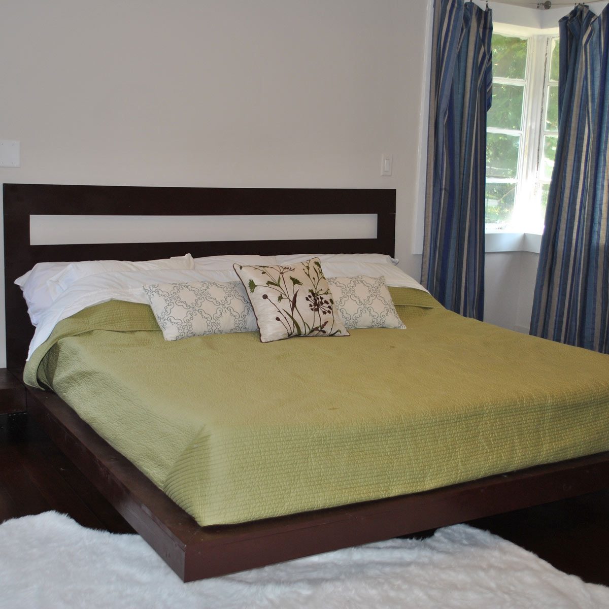 A large bed with a green cover and decorative pillows sits in a bright room, flanked by striped curtains and a window.