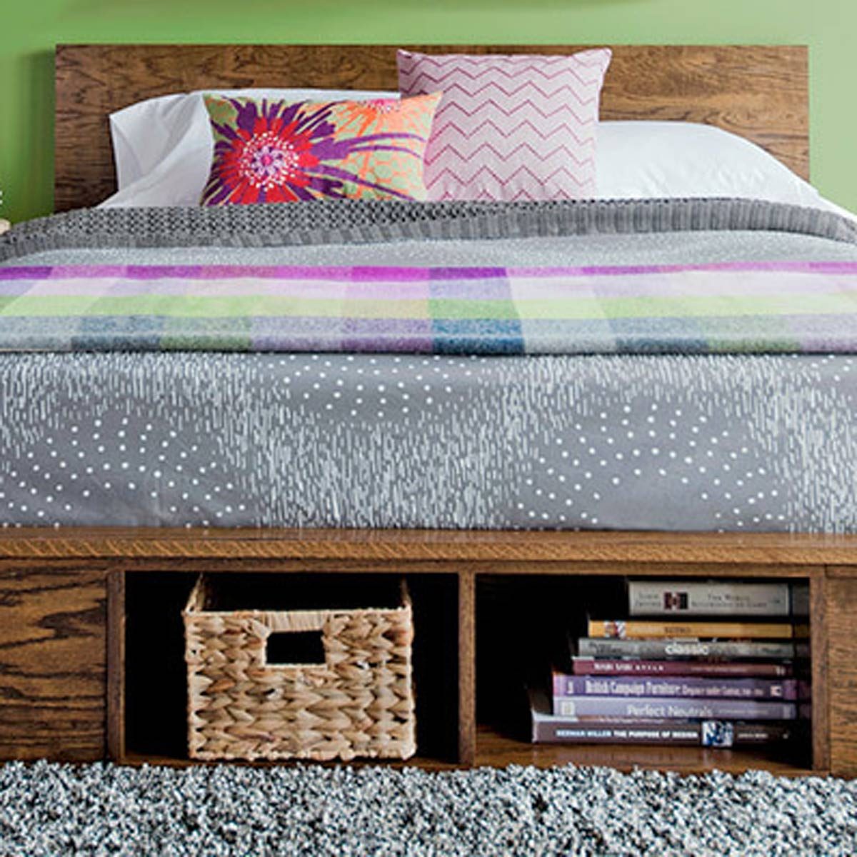 A wooden bed features a colorful quilt and pillows. Beneath, a woven basket and stacked books sit on a plush rug against a green wall.
