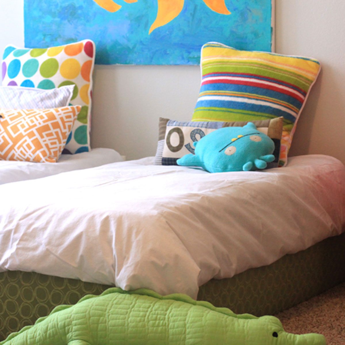 Two beds with colorful pillows and a large green crocodile toy on the floor, in a bright, cheerful room with a blue wall mural.
