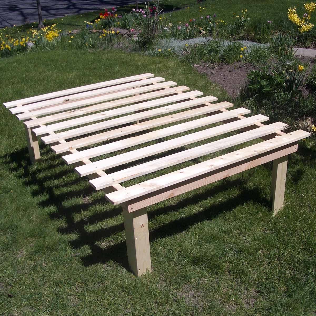 A wooden bed frame rests on green grass, constructed with slats and four sturdy legs, surrounded by colorful flowers and a garden path.