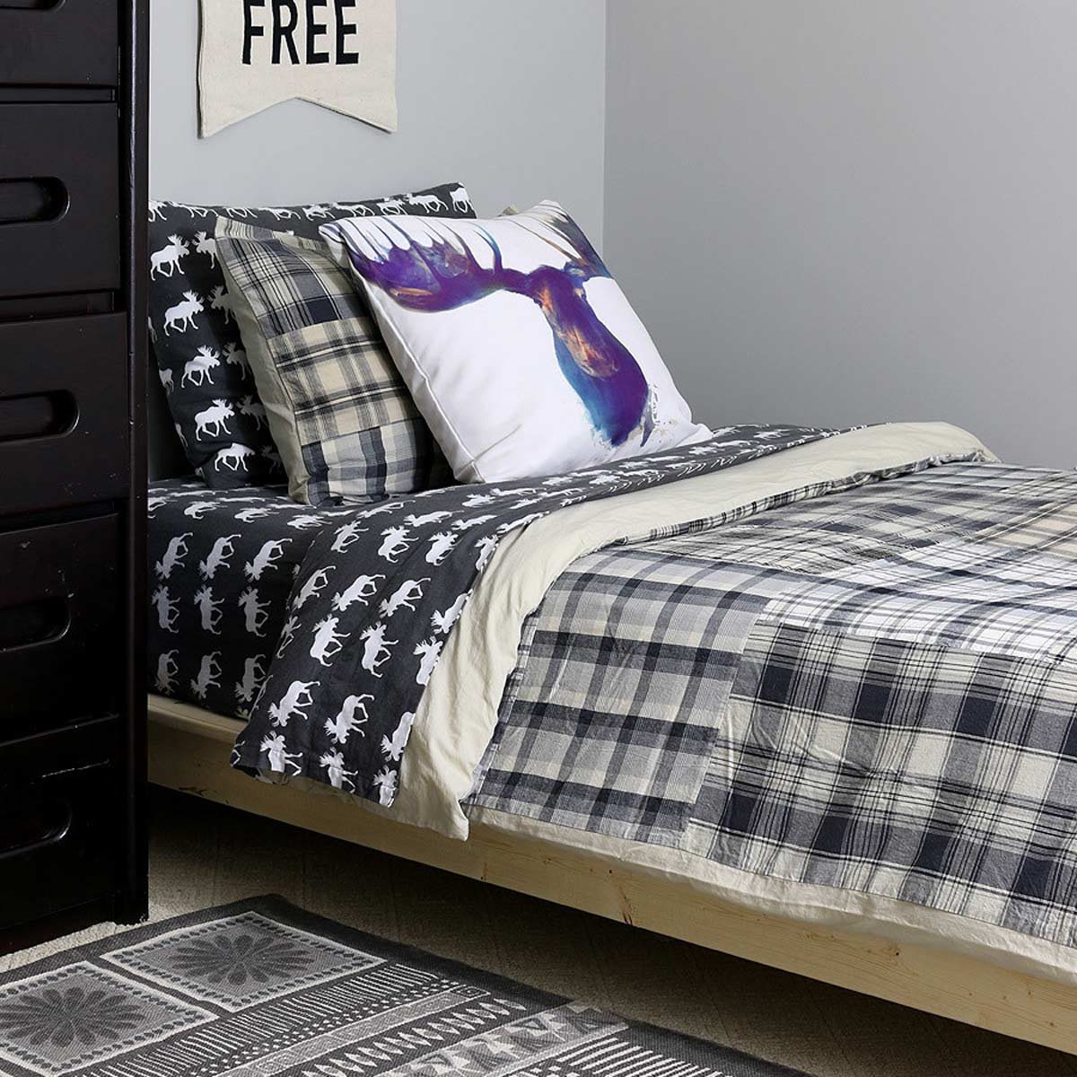 A bed with patterned bedding and pillows displays a moose design, situated against a gray wall and next to a black dresser in a cozy room.