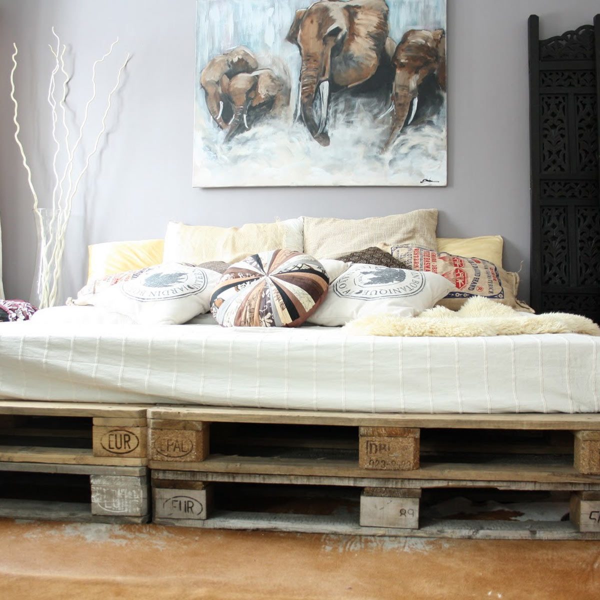 A wooden pallet bed with decorative pillows rests against a wall featuring an elephant painting, surrounded by minimalistic decor and soft lighting.