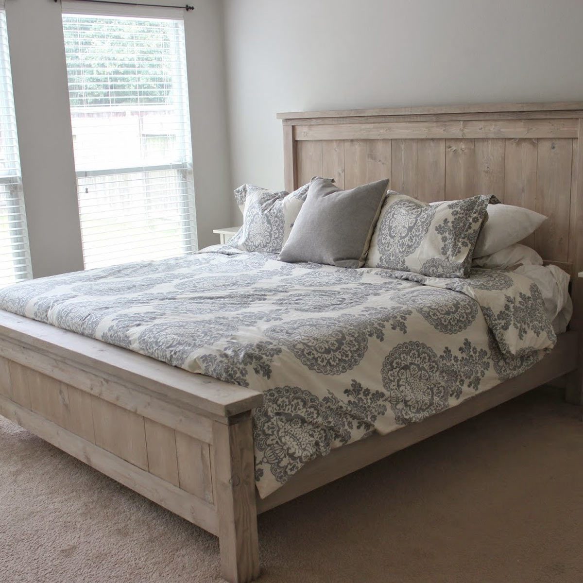 A large wooden bed with patterned bedding and pillows is centered in a room with two windows, allowing natural light to illuminate the space.