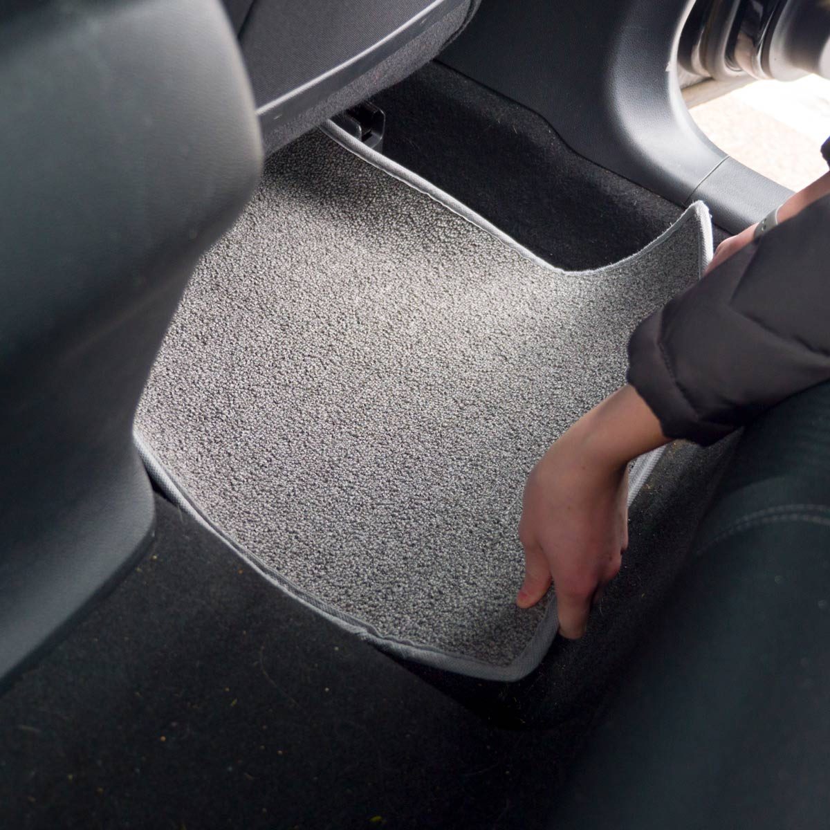 A person is placing a grey car mat into the footwell of a vehicle, surrounded by dark interior elements.