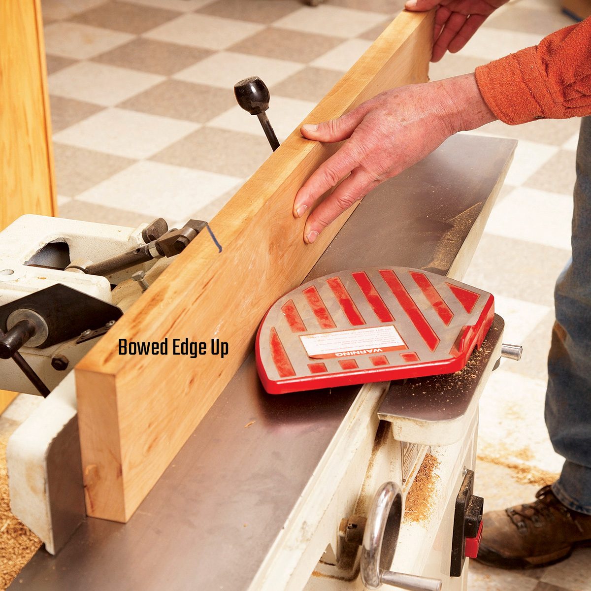 A person guides a wooden board through a jointer, ensuring the bowed edge faces up, in a workshop with a checkered floor.