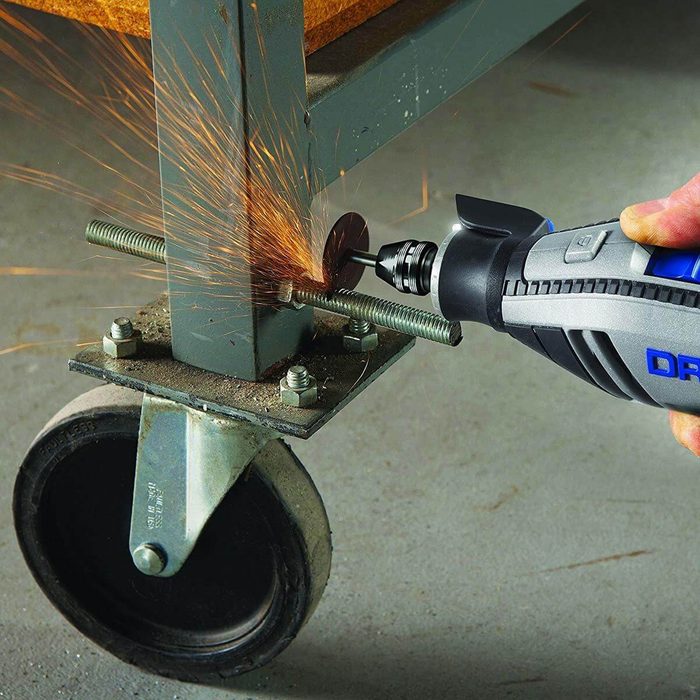 A hand uses a rotary tool to cut metal, producing sparks. The setting features a metal cart with a wheel on a concrete surface.