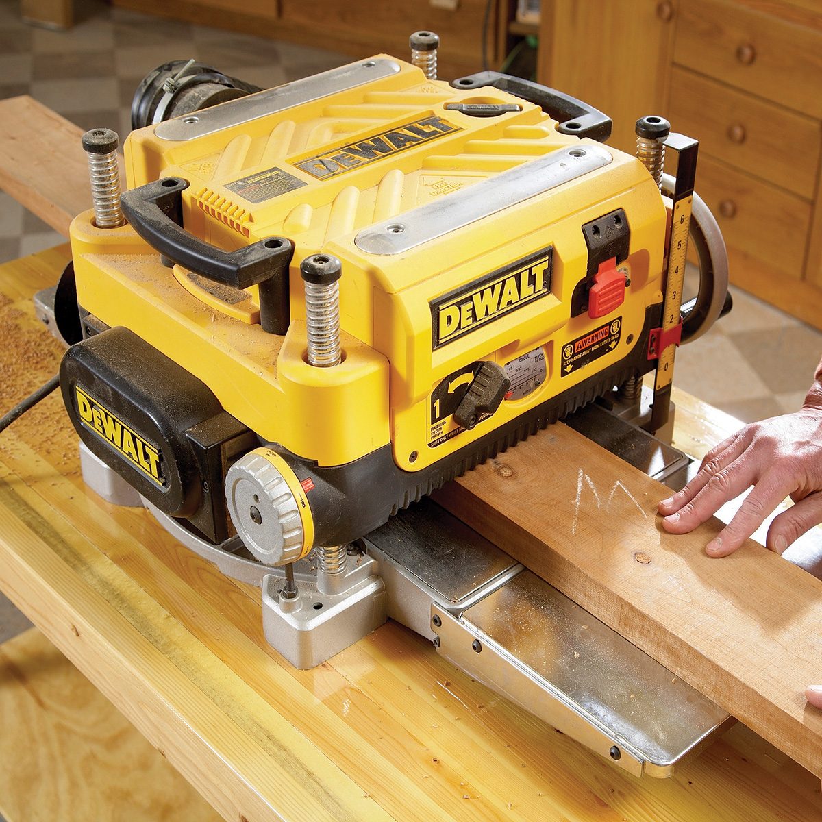 A yellow DEWALT planer smooths a piece of wood while a hand guides it on a wooden workbench, surrounded by a workshop environment.