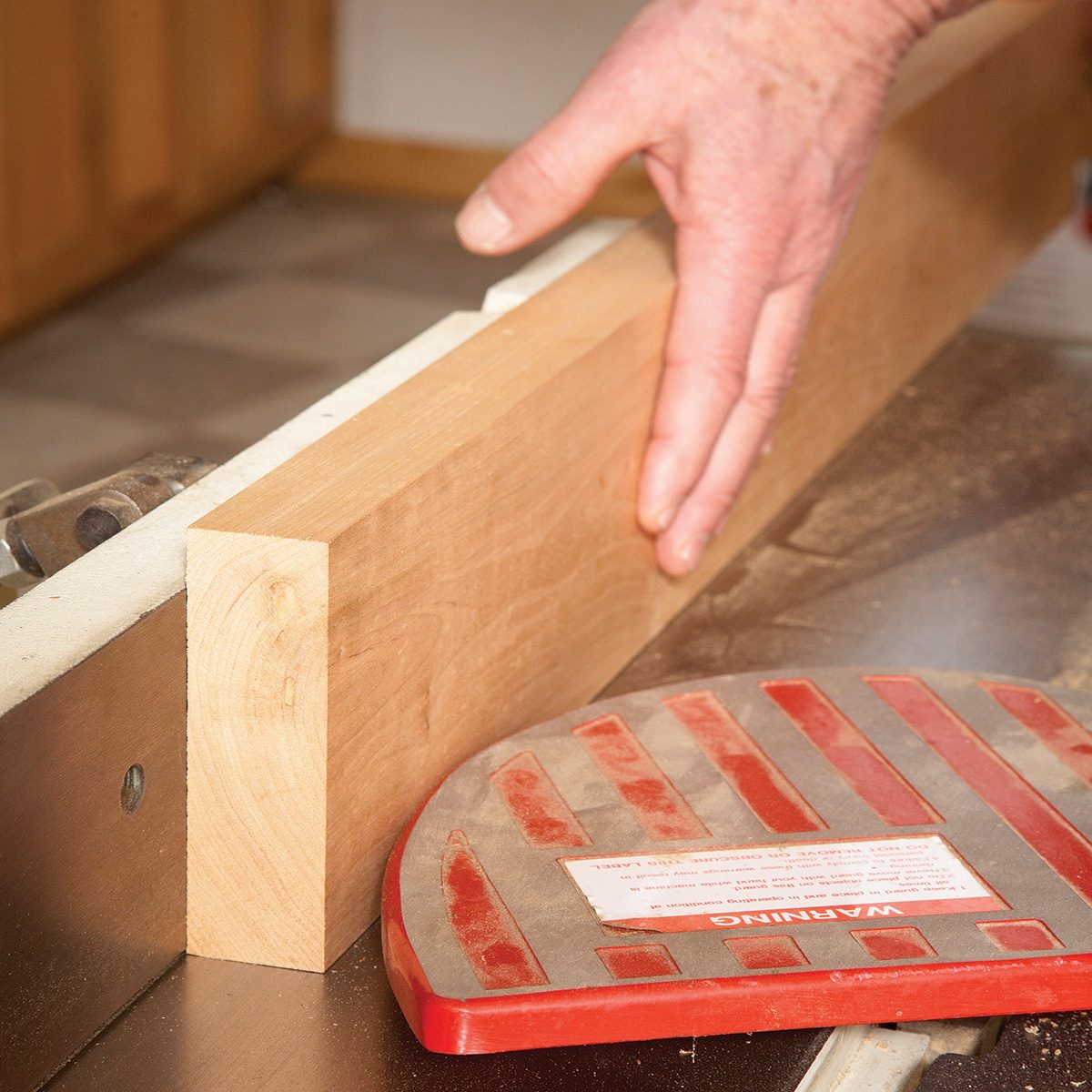 A hand guides a wooden plank along a table saw, ensuring precision while a safety guard is positioned nearby in a workshop environment.