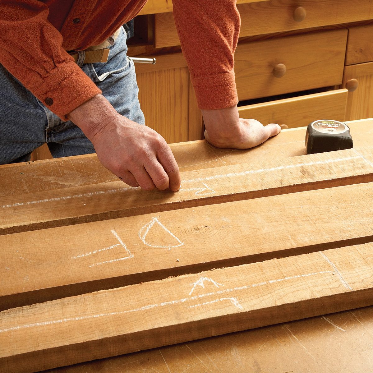 A hand draws measurements with chalk on wooden planks resting on a workbench, surrounded by a well-lit workshop with wooden storage.