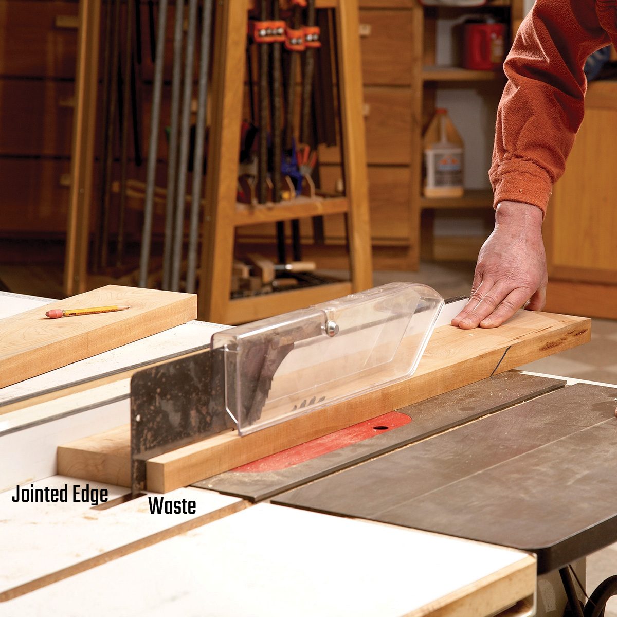 A hand holds a piece of wood against a table saw blade in a workshop, while tools and wooden items are organized in the background.