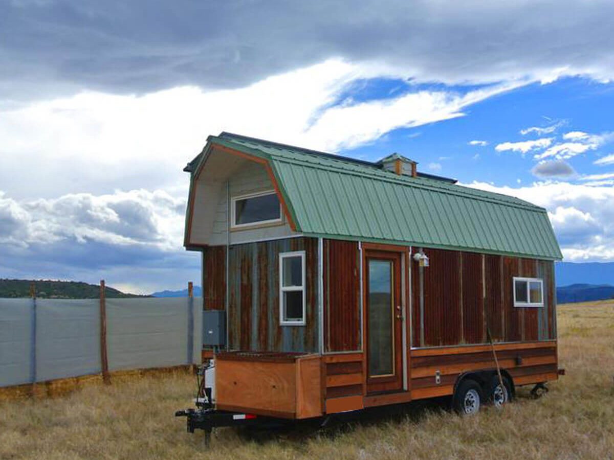 A tiny house on wheels sits in a grassy field under a cloudy sky, showcasing a wooden exterior with a green metal roof.