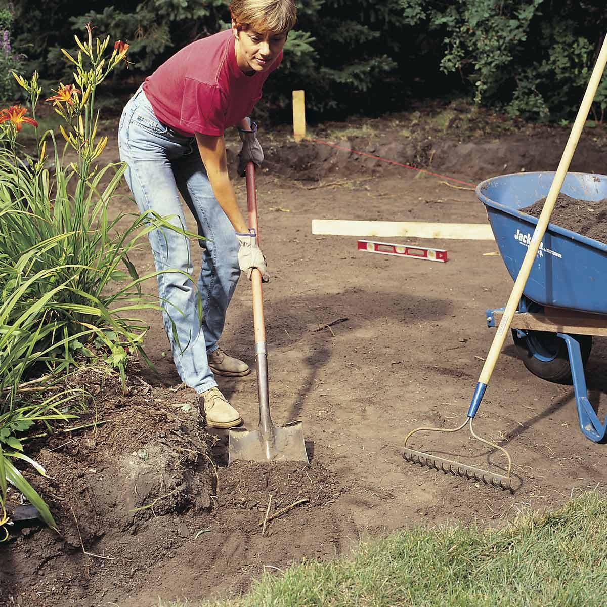 A person digs with a shovel in a garden area, surrounded by plants, while a wheelbarrow and rake are nearby on bare soil.