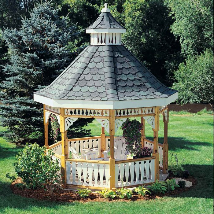 A wooden gazebo with a shingled roof stands in a green yard, surrounded by trees and decorative plants, offering a serene outdoor seating area.