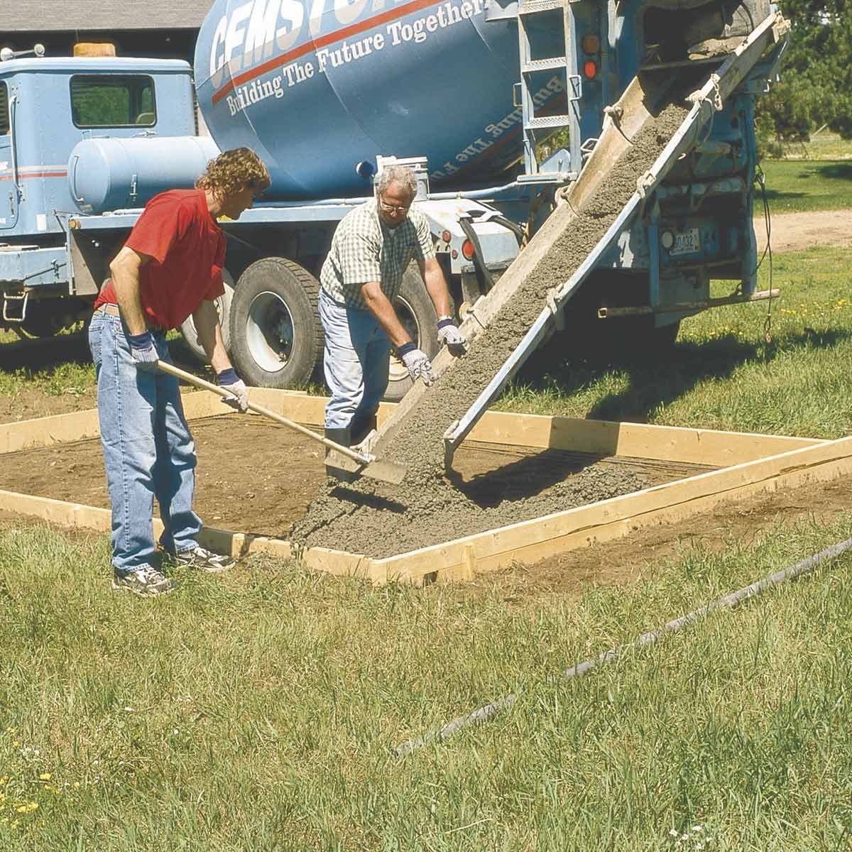 Two men pour concrete into a wooden frame, using shovels, while a cement truck is parked nearby in a grassy outdoor setting.