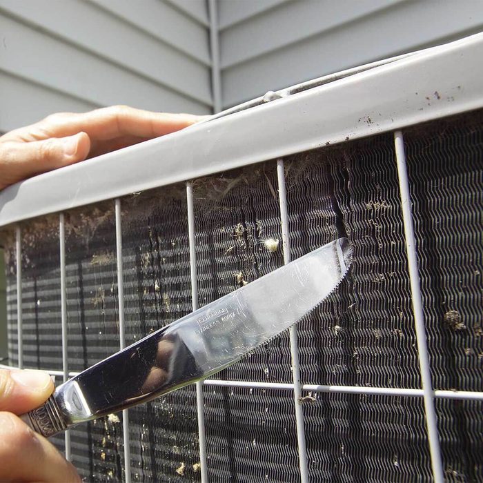 A hand holds a knife, scraping grime from an air conditioning unit's filter. The background shows a house's siding, indicating outdoor maintenance.