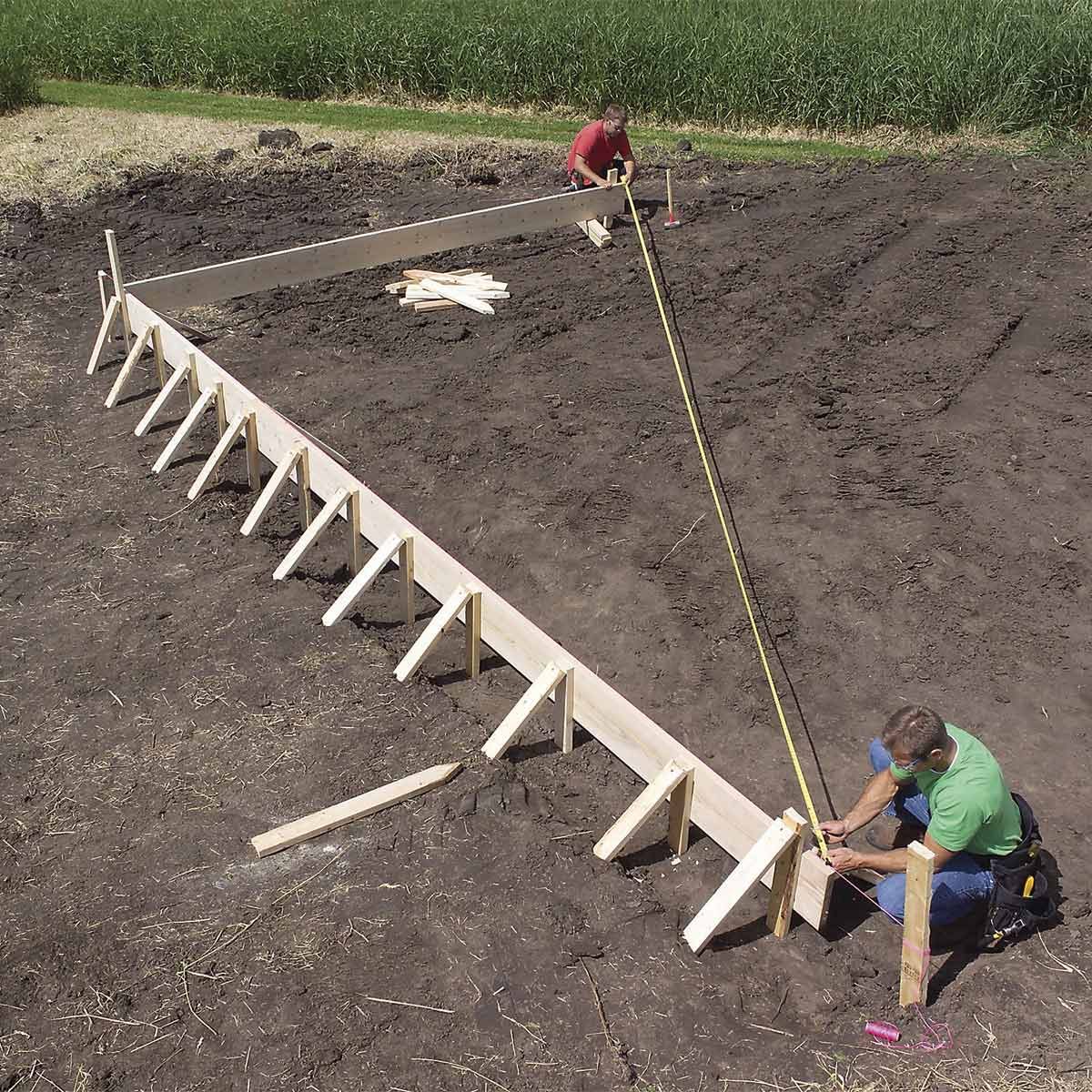Two individuals are constructing a wooden frame on a dirt surface, ensuring proper alignment with measuring tools, surrounded by greenery and grass.