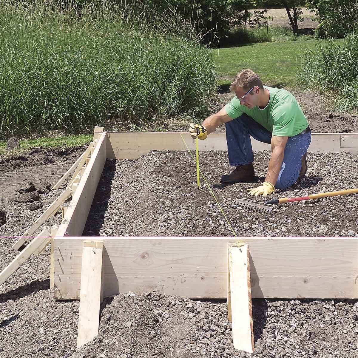 A man kneels on gravel, measuring soil depth with a tape measure. He’s using a rake nearby, surrounded by tall grass and trees.