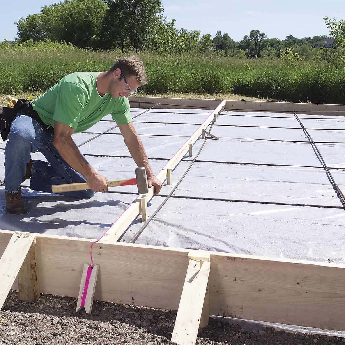 A man kneels, hammering wooden boards on a gravel surface, preparing a foundation with rebar visible underneath, surrounded by green fields and trees.