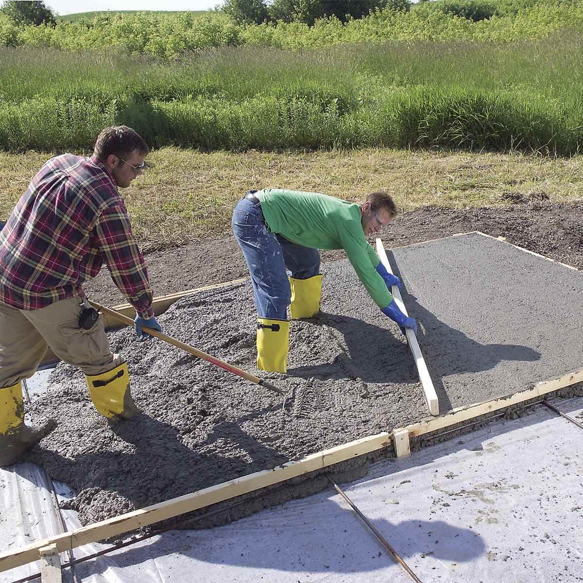 Two workers in rubber boots spread concrete using a board, with grass and trees in the background on a sunny day.