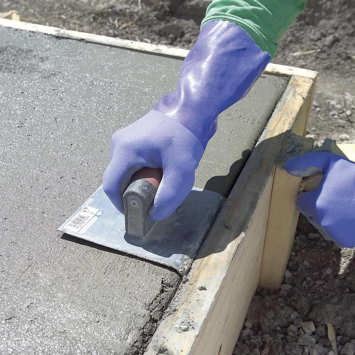 A gloved hand smooths freshly poured concrete with a trowel, shaping the surface alongside wooden forms in a construction environment.