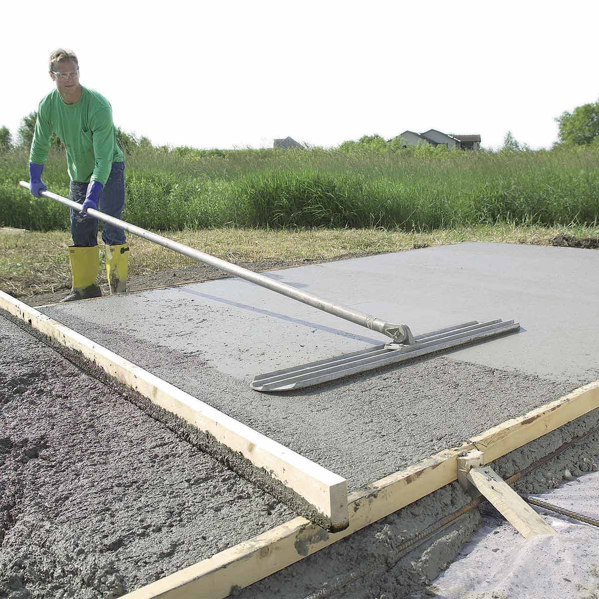 A man in yellow boots levels wet concrete using a long tool while standing on a grassy area beside a wooden frame. Houses are visible in the background.