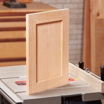 A wooden cabinet door stands vertically on a saw table, showcasing a simple frame design against a workshop background of organized tools and wooden materials.