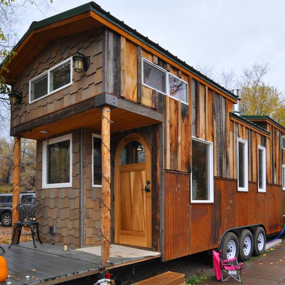 Tiny house stands on wheels, featuring a wooden exterior and large windows. It has a welcoming arched door and is set in a natural environment.
