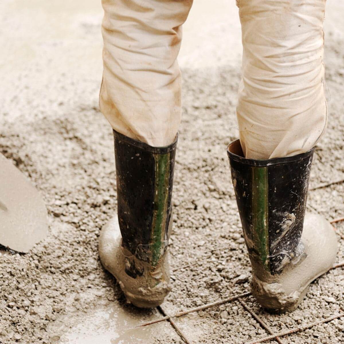 A person in dirty, knee-high rubber boots stands on wet concrete, surrounded by loose gravel, as they work, potentially preparing a surface or mixing materials.