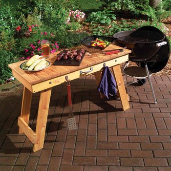 A wooden table displays grilled meats and corn, with a drink nearby, while a charcoal grill sits beside it in a garden setting.