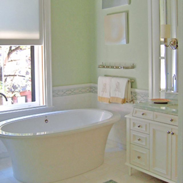 A white bathtub rests in a light green bathroom, beside a vanity with drawers and a sink, while towels hang above a decorative border.