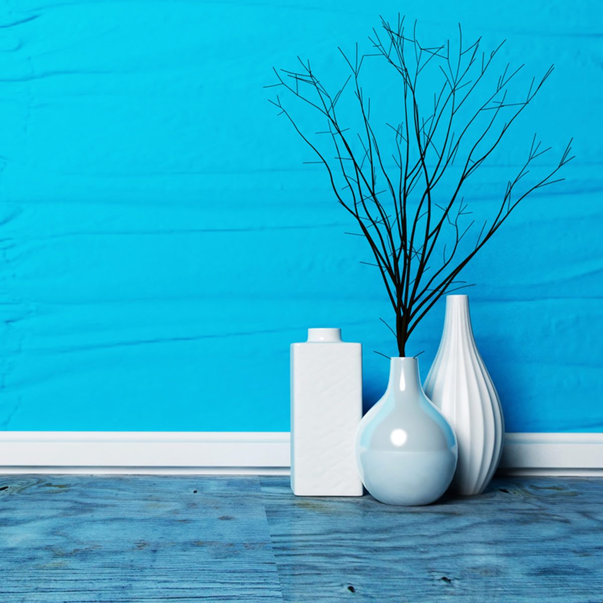 Three white vases display dried branches against a bright blue wall, set on a wooden floor with a textured surface.