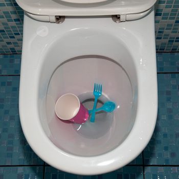 A toilet bowl contains a pink cup, a blue fork, and a blue spoon, surrounded by blue tiled flooring in a bathroom setting.