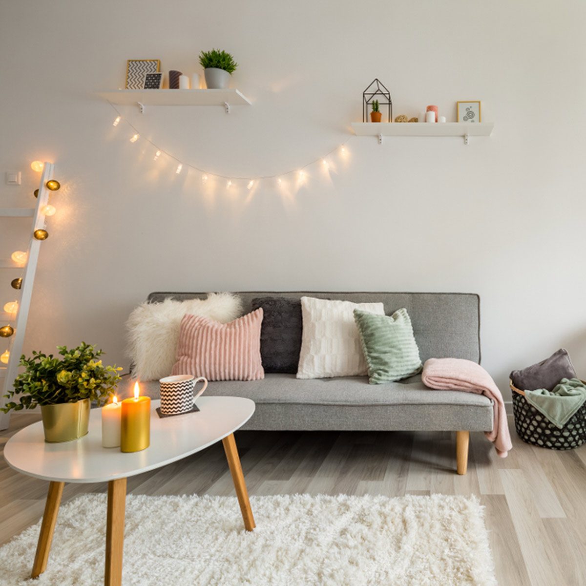 A cozy living room features a gray sofa with colorful pillows, a white coffee table with candles, and warm lights adorning the walls, creating a relaxed atmosphere.