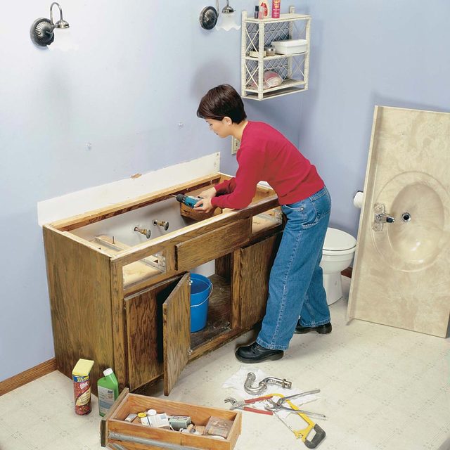 A person installs a sink in a bathroom, surrounded by tools and materials, with an open cabinet and a toilet in the background.