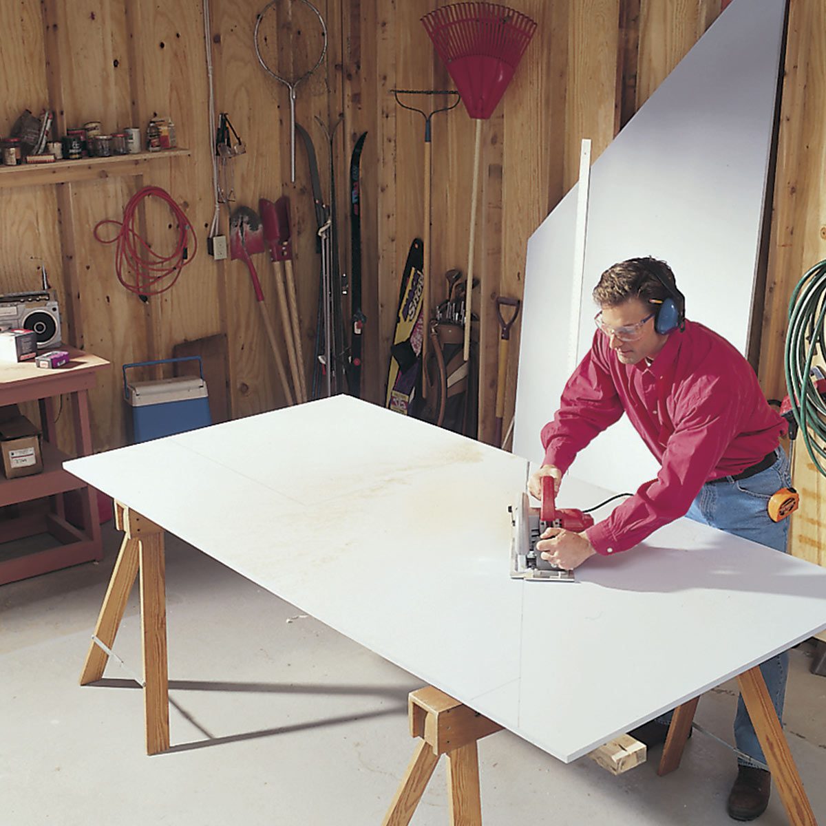 A man uses a circular saw on a large white board positioned on sawhorses, inside a workshop filled with tools and equipment.