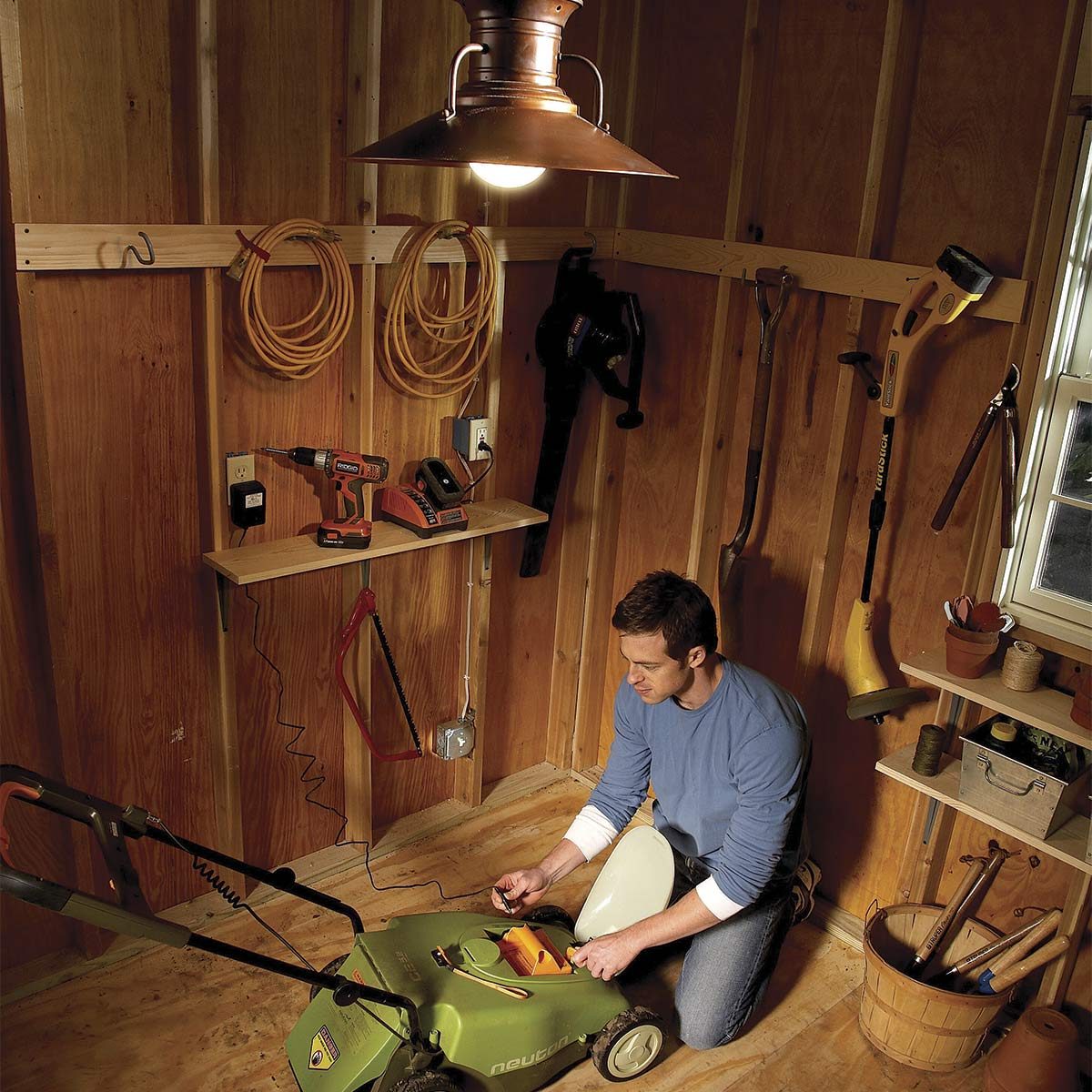 A man is kneeling beside an electric lawn mower, connected to a power source, in a wooden shed filled with tools and gardening supplies.