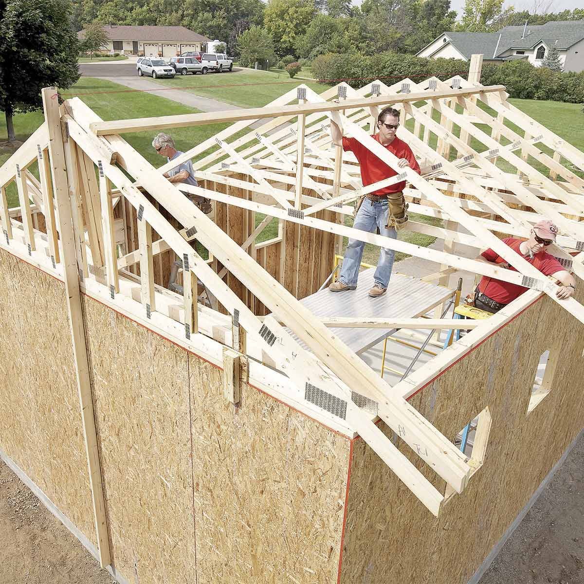 Workers are constructing a wooden roof frame on a house, surrounded by grassy lawns and residential buildings, with scaffolding visible for support.