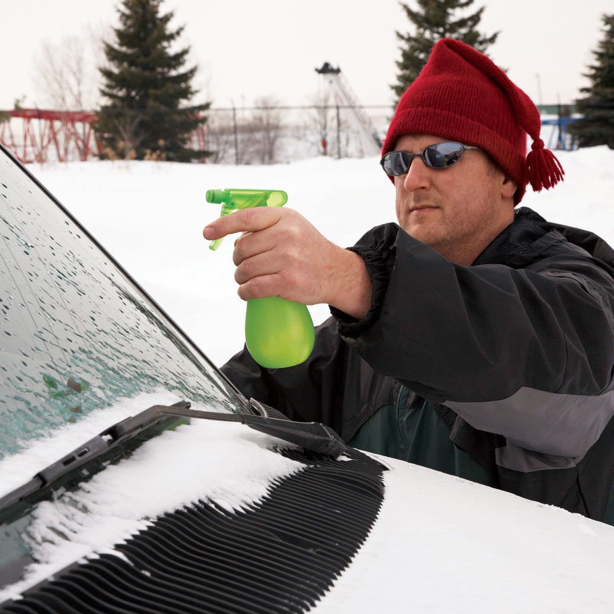 A man sprays a green liquid onto a snowy car windshield, preparing to clear it, in a cold, snowy environment with evergreen trees nearby.