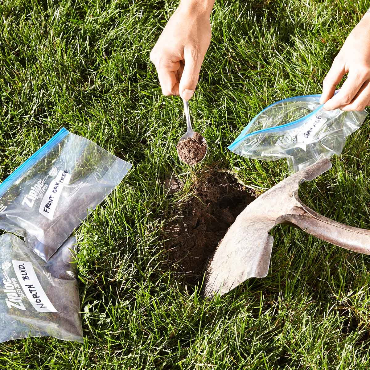 A hand scoops soil with a spoon, placing it into a labeled plastic bag beside an open hole and a gardening tool, on grassy terrain.