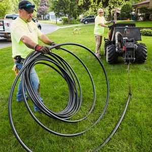 Two workers are laying black tubing in a grassy yard using a machine, while one employee manages the coiled tubing. A sunny suburban environment surrounds them.